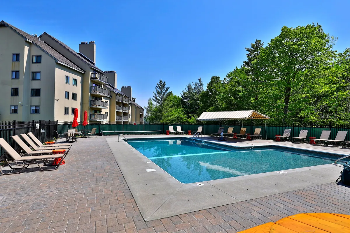 An inviting outdoor swimming pool surrounded by lounge chairs, lush green trees, and the building's facade. The pool area is sunlit and designed for summer leisure and gatherings.
