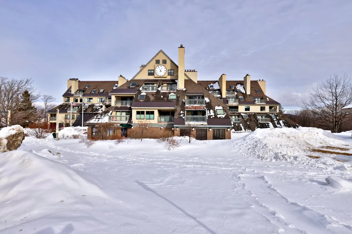 A snow-covered building with a prominent clock tower, featuring balconies and surrounded by trees. The rooftops and landscape are blanketed in white, evoking classic winter mountain resort vibes.