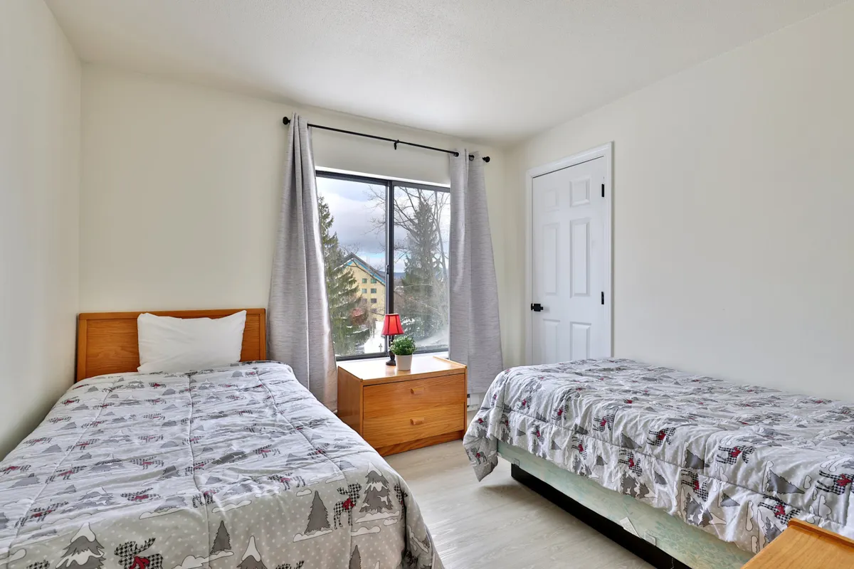 A serene second bedroom with two twin beds, a large window letting in natural light, and a nightstand with a lamp, creating a cozy and inviting space ideal for children or friends. The overall look is both bright and comforting, perfect for restful nights.
