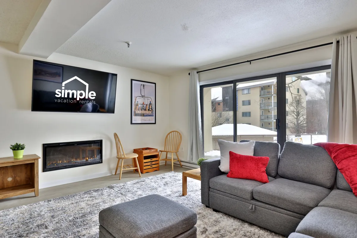 A cozy, open living room featuring a comfortable gray sofa positioned near a modern electric fireplace. Large windows fill the space with natural light, revealing outdoor winter scenery. A flat-screen TV sits above the fireplace, and the seating area is designed for relaxation, conversation, or movie nights.