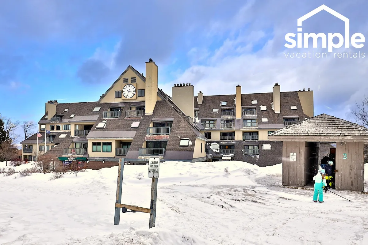 An exterior view of the Mountain Green building featuring a prominent clock tower, snow-covered ground, and the main entrance structure. The surrounding environment suggests easy access to winter sports and après-ski activities.