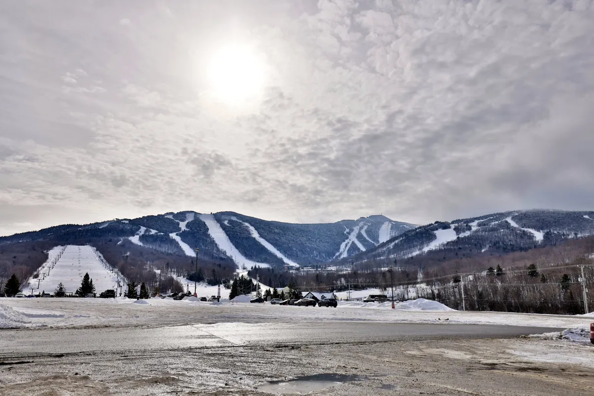 A panoramic outdoor view of snow-covered mountains and ski slopes, bathed in sunlight, with visible ski runs and traces of skiers and snowboarders. The scene captures the excitement and adventure of being right at the heart of Killington’s winter sports action.
