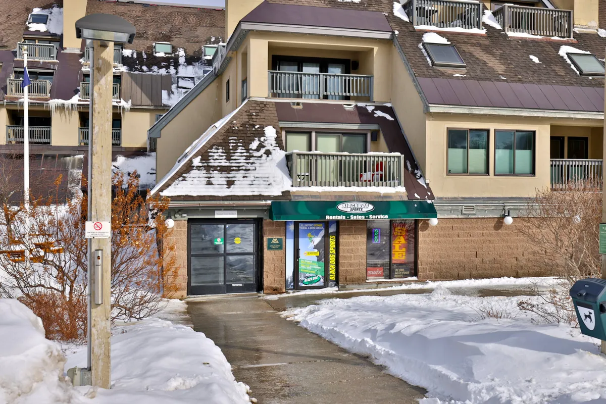 A welcoming exterior shot of the Mountain Green entrance with a green awning, snow-covered pathways, and the building's facade featuring balconies. The scene is lively and hints at winter activities, with spaces for groups to gather after a day on the mountain.