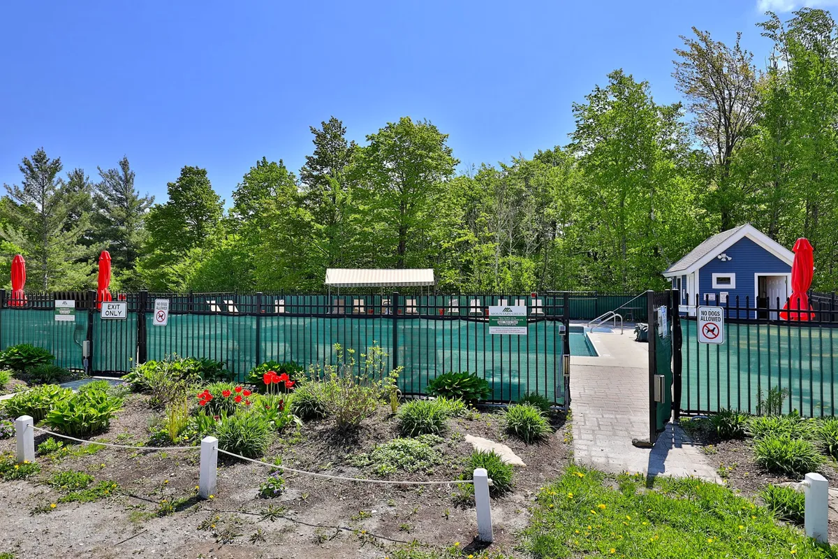 A sunny exterior image of a vibrant outdoor swimming pool bordered by colorful umbrellas and landscaped gardens, with families and friends enjoying the water and lounging poolside. The pool house and green spaces highlight summer activities and relaxation.