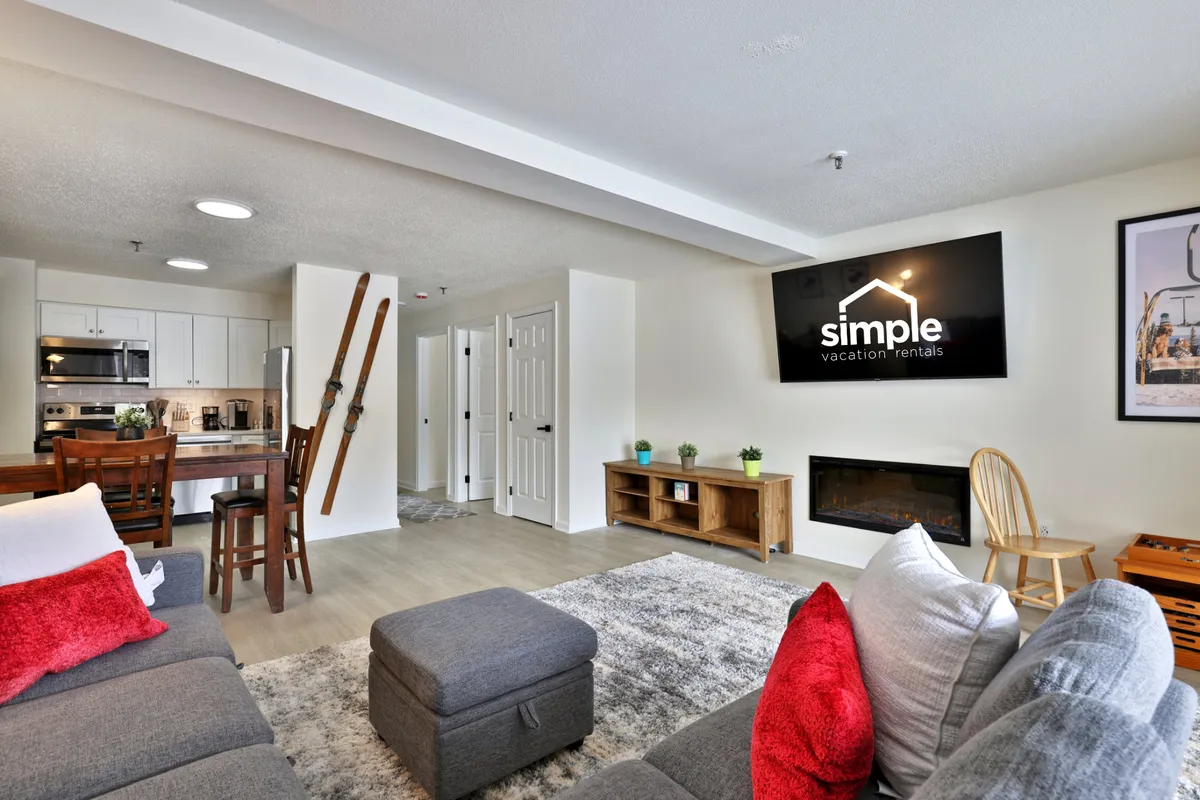 A comfortable interior with a sofa decorated in red and white pillows, a wall-mounted TV, a fireplace, and a dining area with wooden chairs. The setup suggests a perfect blend of relaxation, entertainment, and warmth.
