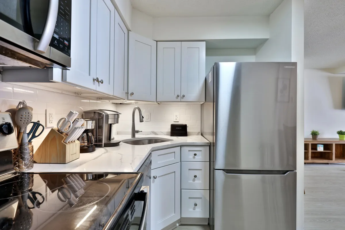 This image showcases a functional kitchen with a stainless steel refrigerator, modern oven, kitchen sink, coffee maker, and a cutting board ready for meal prep. The setup is cozy yet highly practical, reflecting a space well-equipped for cooking, socializing over coffee, or preparing snacks.
