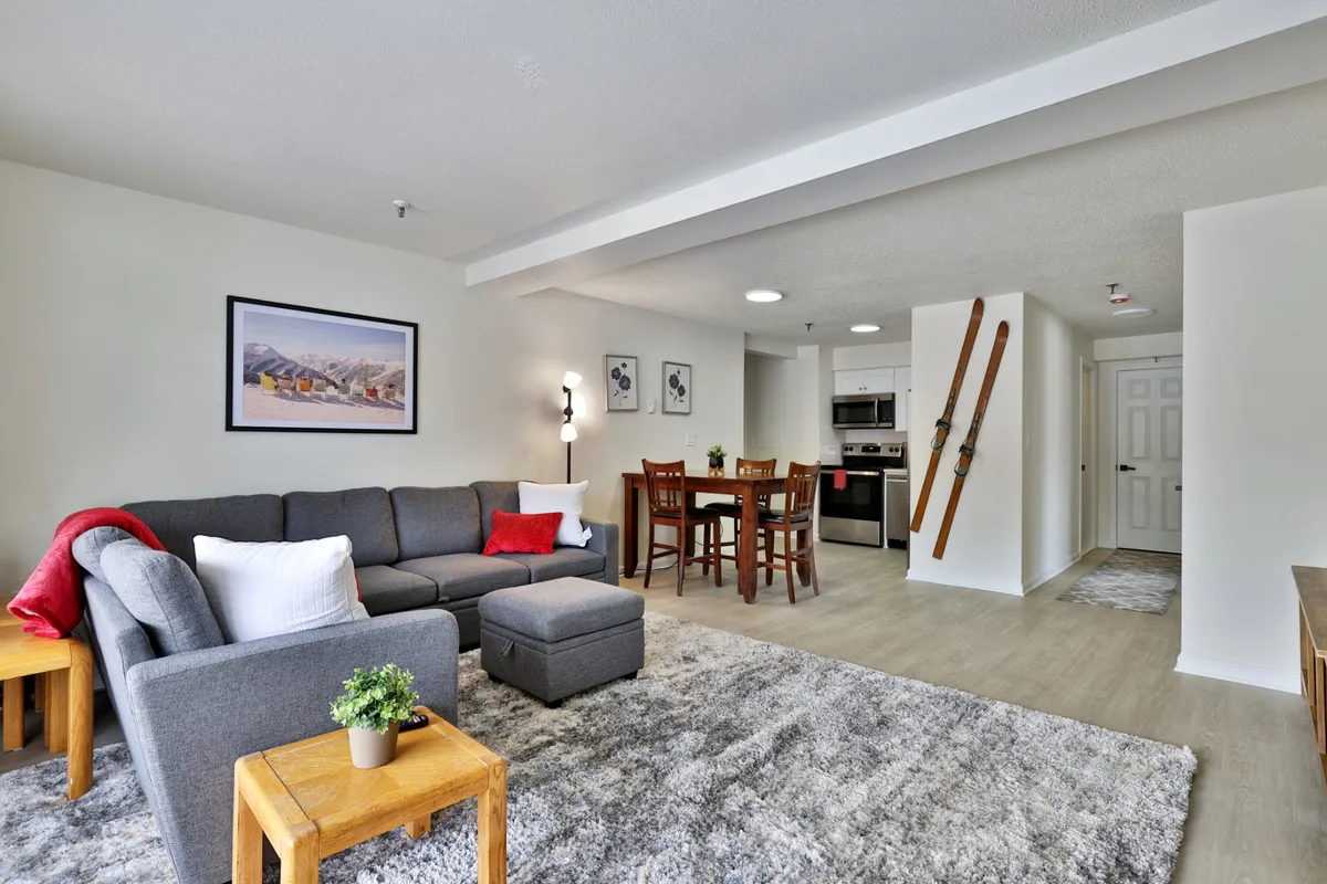 The image showcases a stylishly renovated open-concept living space featuring a plush sofa, inviting dining table, modern kitchen, and tasteful ski-themed decor. Warm lighting and cozy accents create a welcoming atmosphere perfect for relaxation and social gatherings after a day on the slopes.